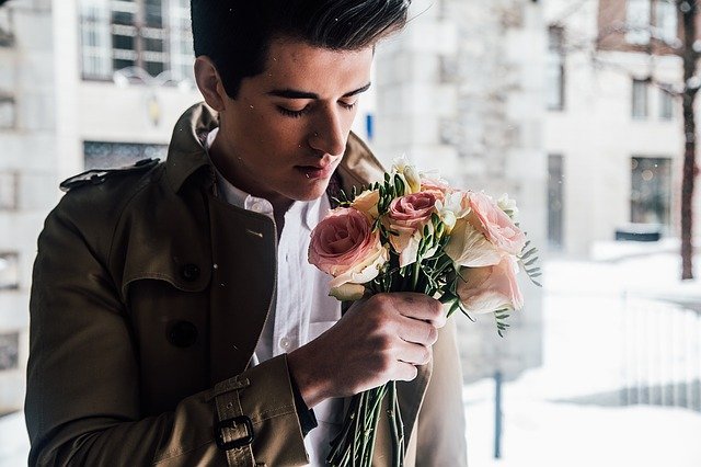 白とくすんだピンク色の薔薇の花束をもつ、ベージュのミリタリーコートを着た欧米人の若い男性の写真。