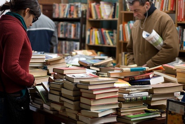 欧米の古書店で積み上げられた本の山を見下ろすサングラスの若い女性と、イアホンをした中年の男性の写真。