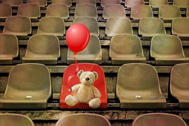 薄い茶色の座席の無観客スタジアムに、赤い風船を持ったテディベアが、１つだけある赤い席に座っている。