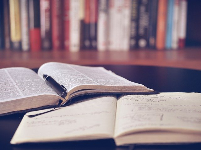 ピンボケした本の並びを背景に、机上で広げられたノートの上に、ボールペンが乗った細かい英字のびっしり記された本が開かれている写真。