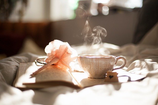 朝の陽光の射す部屋でノートに置かれた湯気の出るコーヒーカップのあるオシャレな部屋の写真。