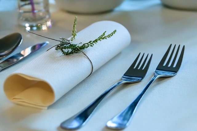 ホテルの、白いクロスのテーブルに置かれたカトラリー（ナイフ、フォーク、スプーン）と、植物で結ばれたナプキンの写真